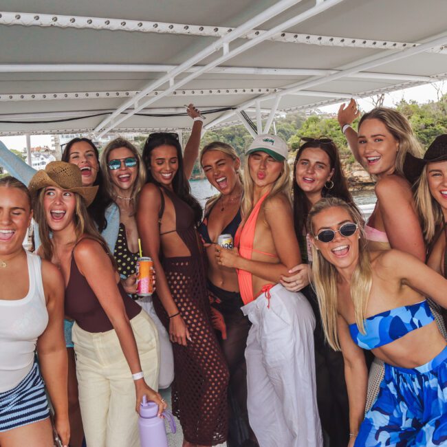 A group of smiling women in swimwear pose together on a boat, celebrating and enjoying a sunny day on the water, with trees and buildings visible in the background.