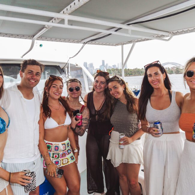 A group of eight smiling people, dressed in swimsuits and summer clothing, stand together on a boat holding drinks. The background shows water, trees, and a city skyline under a sunny sky.