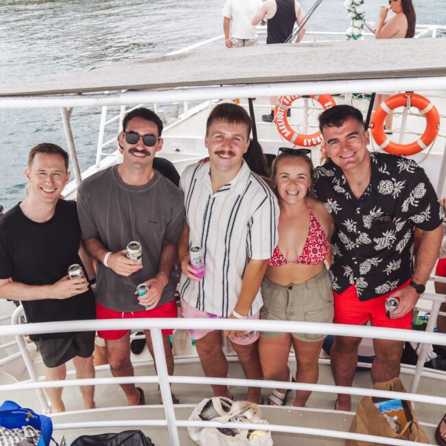 Five people stand close together and smile at the camera on a boat deck, holding drinks. They are casually dressed for warm weather, with water, life rings, and other people visible in the background.