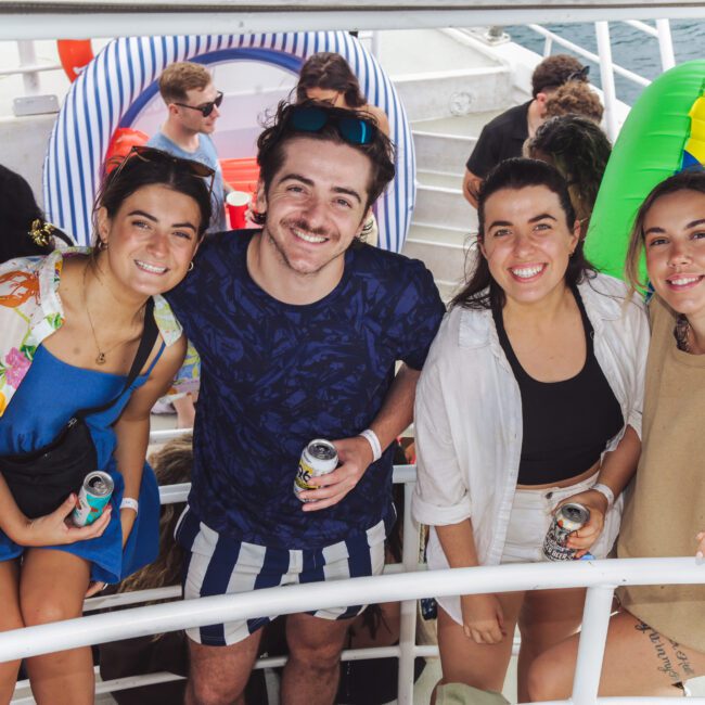 Four young adults smile at the camera while holding drinks on a crowded boat. Colorful pool floats and the ocean are visible in the background, suggesting a lively summer party atmosphere.