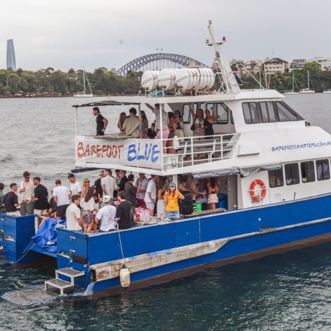 A blue and white double-decker boat called "Barefoot Blue" sails on the water with a large group of people socializing and enjoying a party; city buildings and greenery appear in the background.