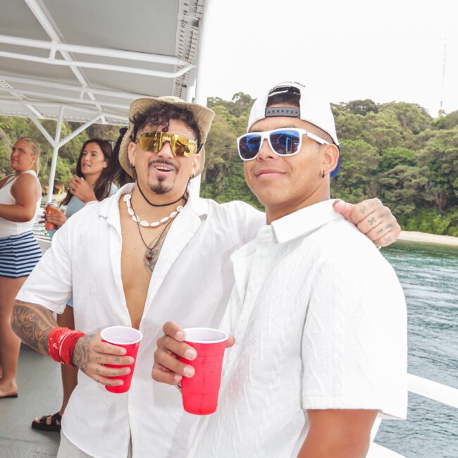 Two men in white shirts and sunglasses smile on a boat, each holding a red cup. Other people stand in the background, with water and greenery visible outside. The atmosphere appears relaxed and festive.