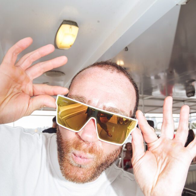 A man with a beard and large, reflective sunglasses poses playfully, holding the sunglasses with both hands and puckering his lips. He is indoors under bright lighting, wearing a white shirt.