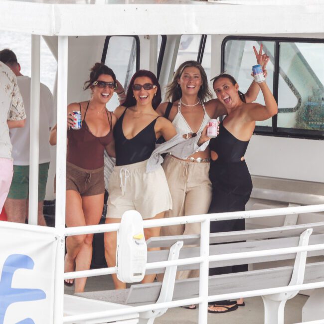 Four smiling women in swimsuits and casual cover-ups stand on a boat deck, holding drinks and posing for the camera; one woman flashes a peace sign. The scene appears cheerful and relaxed.
