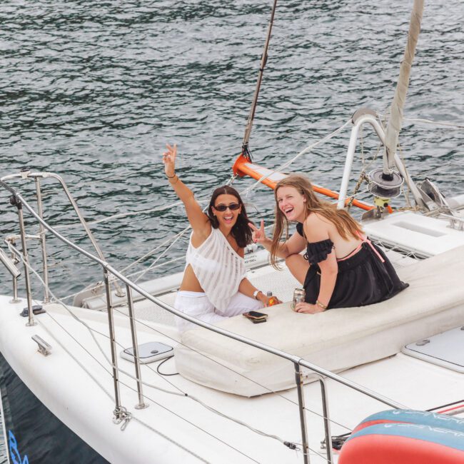 Two women sit and smile on the deck of a sailboat, one waving cheerfully at the camera. They are surrounded by water, with another person relaxing nearby. The scene looks joyful and summery.