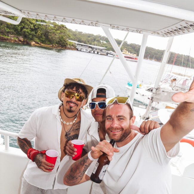 Three men wearing white outfits and sunglasses smile and pose with drinks on a boat. The background shows water, trees, and a dock under a cloudy sky.
