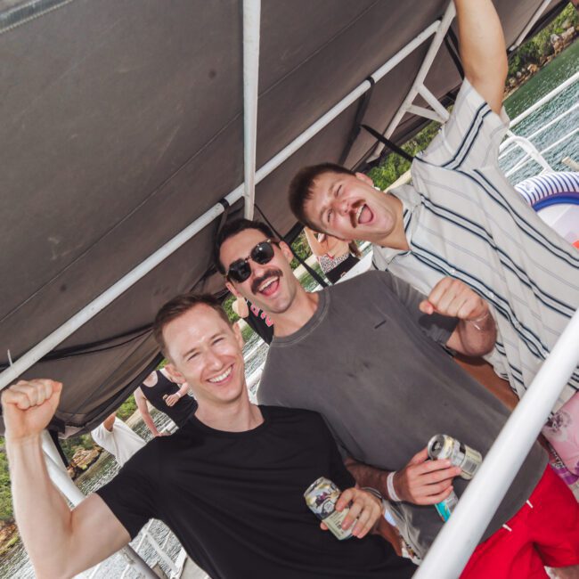 Three men smile and cheer under a canopy on a boat, each holding a drink. There is greenery and water in the background, and other people are partially visible enjoying the scene.