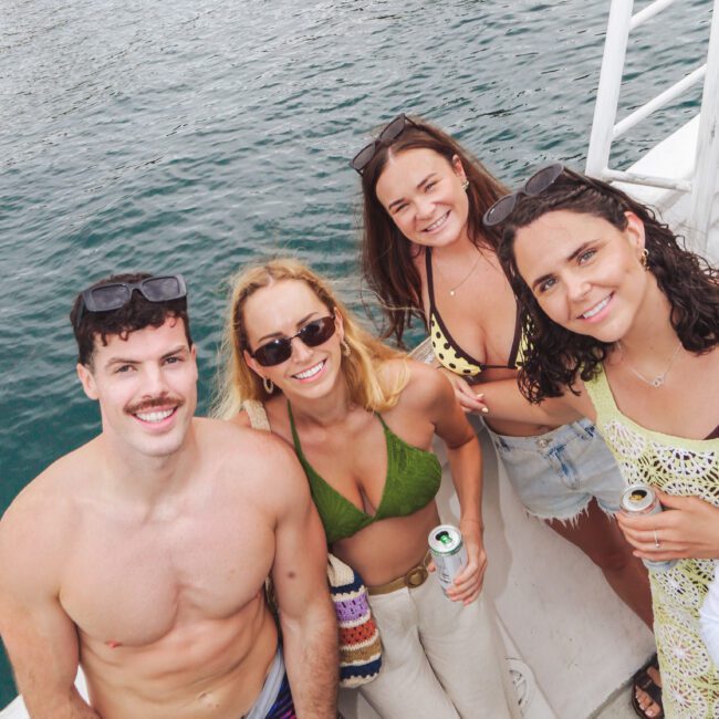 Four people smiling at the camera on a boat, holding drinks. The group includes three women in swimwear and one shirtless man. The water is visible in the background, and they appear to be enjoying a sunny day.