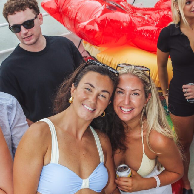 Two smiling women in swimsuits pose together holding drinks on a boat deck, with other people nearby. Behind them is a large inflatable red lips pool float and orange life rings attached to the railing.
