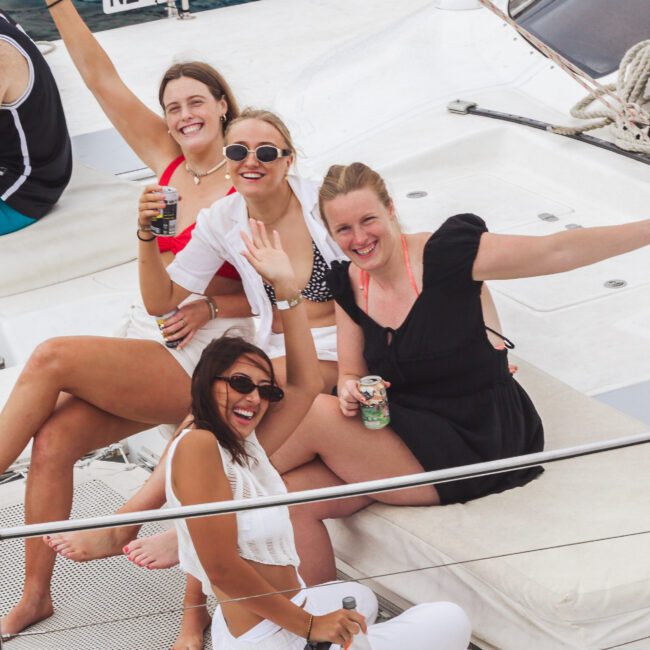 Four women are smiling and waving while relaxing on the deck of a boat. They are wearing swimsuits and summer outfits, holding drinks, and enjoying a sunny day near the water.