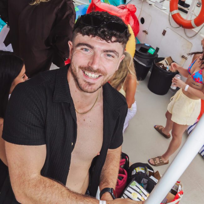 A man with short dark curly hair and a trimmed beard smiles at the camera while holding a drink on a boat. He is wearing a black open shirt and patterned swim trunks. Other people and colorful bags are visible in the background.