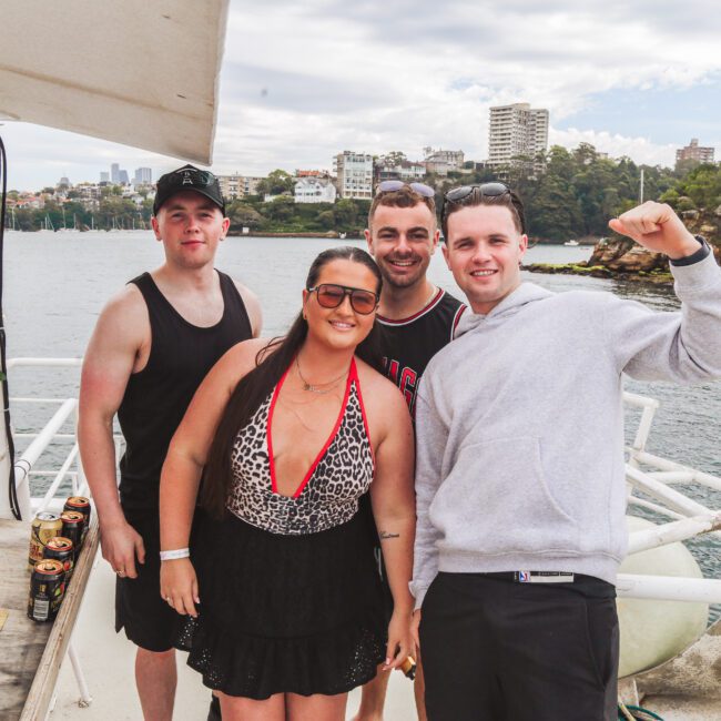 Four people pose and smile on a boat with a cityscape and trees in the background. One man raises his arm in excitement. Cans are visible on a table beside them, and the weather is cloudy.