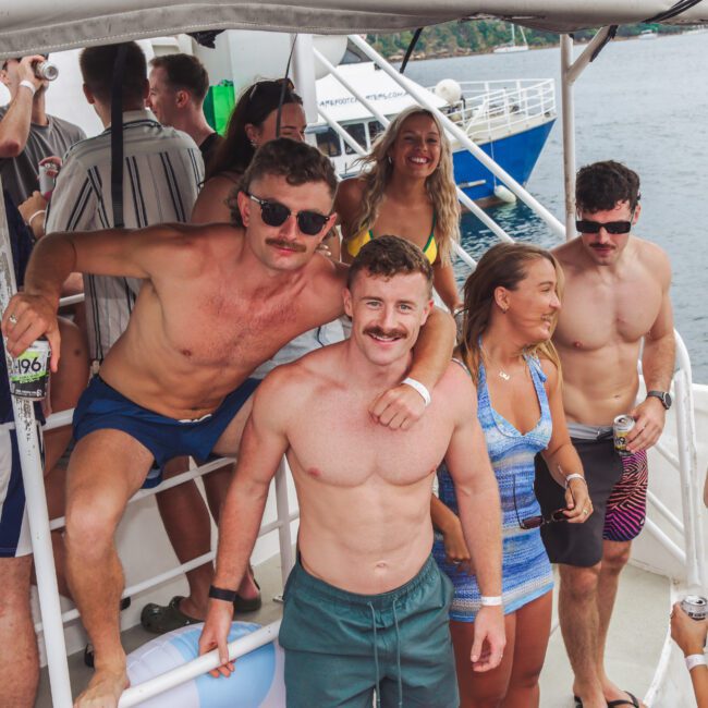 A group of young adults in swimwear, some with mustaches, smiling and posing on a boat. The scene appears festive, with people enjoying themselves by the water on a cloudy day.