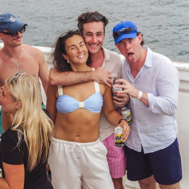 Four young adults on a boat, smiling and posing for a photo. The woman in front wears a light blue bikini top and white pants. The man behind her hugs her, while another man in a blue cap makes a funny face holding a drink.
