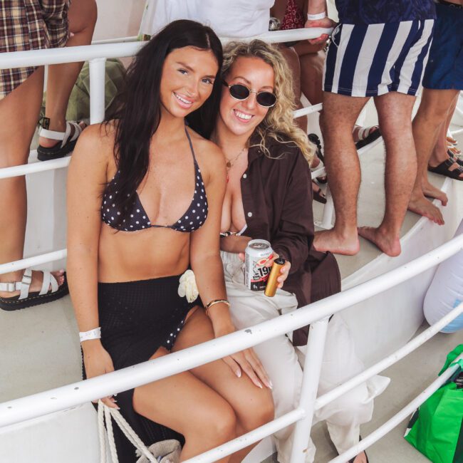 Two women sit and smile on a boat surrounded by other people. One wears a black polka-dot bikini top and black skirt, the other wears sunglasses, a brown shirt, and holds a can of drink. Beach bags are on the floor nearby.