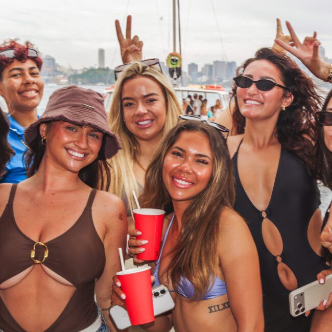 A group of seven young women smiling and holding red cups pose for a photo outdoors. Some make peace signs, and the city skyline is visible in the background on a cloudy day.