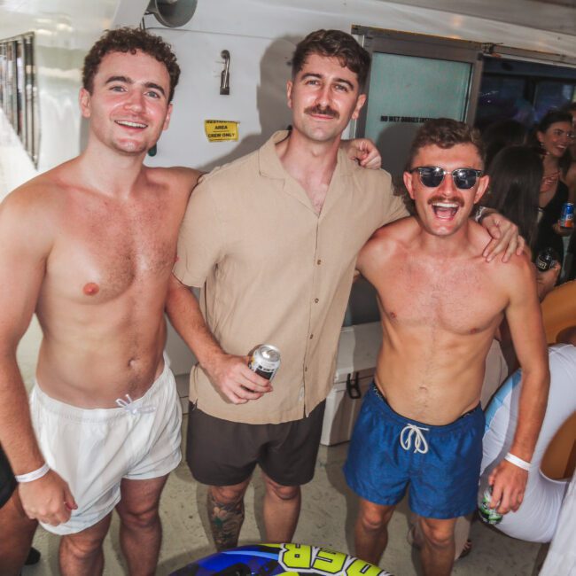 Three young men smile and pose together at a party on a boat. Two are shirtless in swim trunks, and one is wearing a beige shirt. People and colorful pool inflatables are in the background.