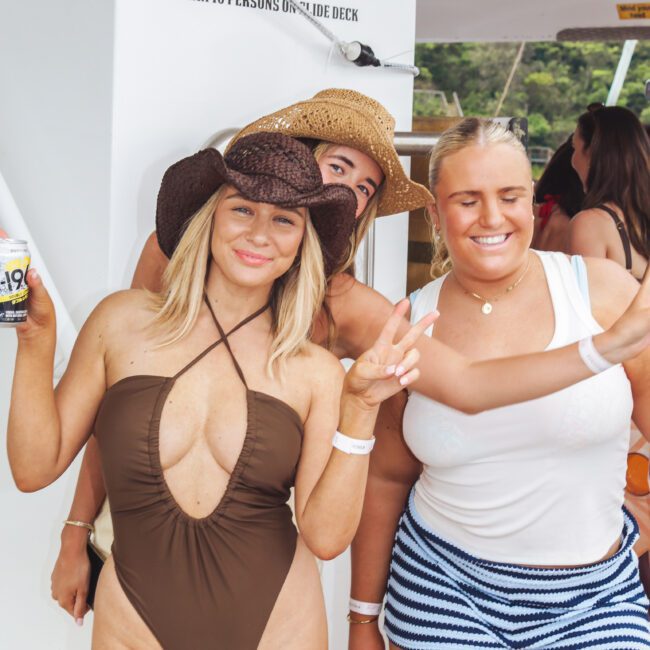 Three women in swimsuits, two wearing brown hats, pose and smile on a boat. One holds a drink and the other flashes a peace sign. There is a “MAX 10 PERSONS ON SLIDE DECK” sign in the background.