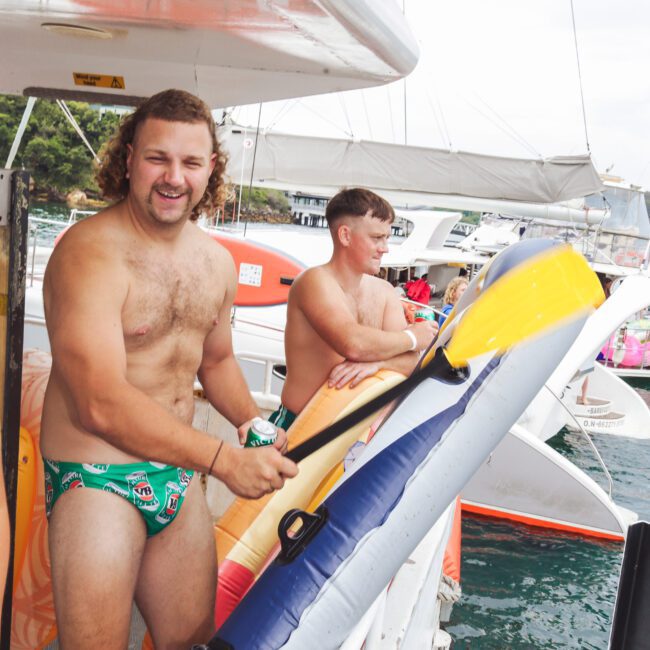 Two shirtless men stand on a boat by the water; one is smiling at the camera wearing patterned swim briefs and holding an inflatable kayak, while the other looks away, also in swim trunks. Other boats and people are in the background.
