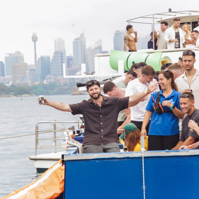 A group of people enjoy a lively boat party on the water with drinks, smiles, and city skyscrapers visible in the background under a bright sky. One man stands at the edge with arms wide open, posing for the camera.