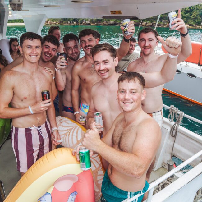 A group of smiling shirtless men holding drinks and inflatables pose together on a boat, with water and another boat visible in the background. The atmosphere appears relaxed and festive.