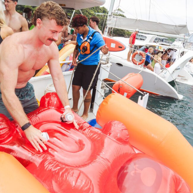 A shirtless man on a boat inflates a large red gummy bear-shaped pool float. Other people stand and sit nearby, with more boats and water visible in the background.
