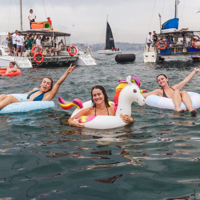 Three women smile and pose on inflatable floats in the water near several boats during a party; two women are on unicorn floats and one on a seashell, with people watching from nearby yachts.