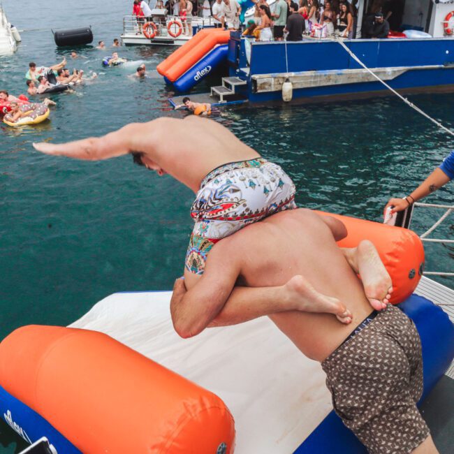 Two men in swim trunks are playfully diving off an inflatable platform into the water, while people swim and relax on floats nearby. A woman stands watching on the ramp, with boats and a crowd in the background.