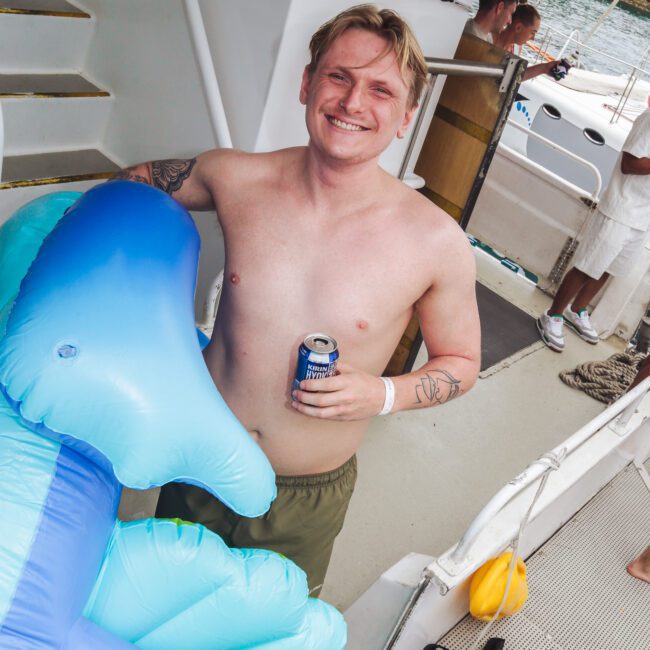 A smiling, shirtless man with tattoos holds a can of beer and stands beside a blue inflatable pool float on a boat. Other people are in the background, and the boat is docked near the water.