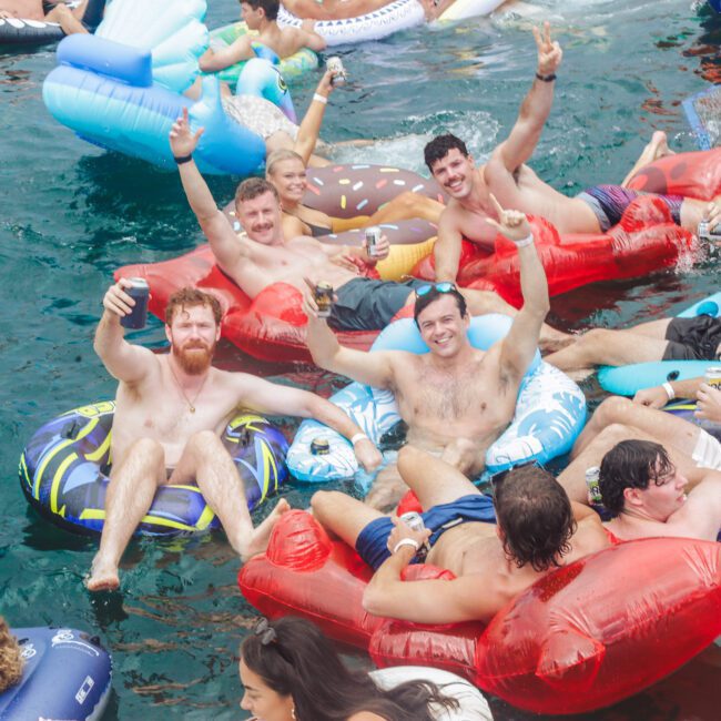 A group of men in swim trunks are floating on colorful inflatable tubes in the water, smiling, raising their arms, and holding drinks, enjoying a lively and festive atmosphere.