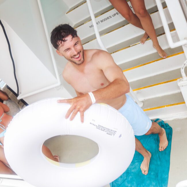 A smiling man in swim trunks stands on a blue towel holding a white inflatable pool ring on a boat. Several other people in swimwear are in the background near stairs. The scene looks lively and summery.