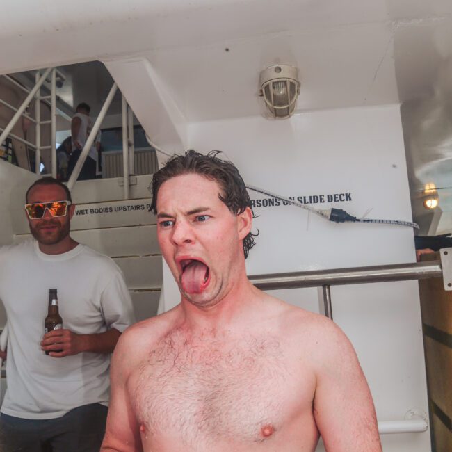 A shirtless man with wet hair makes a funny face, sticking out his tongue and squinting. Another man in sunglasses and holding a beer stands in the background on a boat with white railings and stairs.
