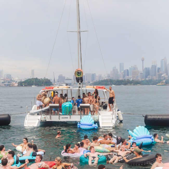 A group of people on a catamaran and in the water on colorful inflatables, enjoying a party. The city skyline and cloudy sky are visible in the background.