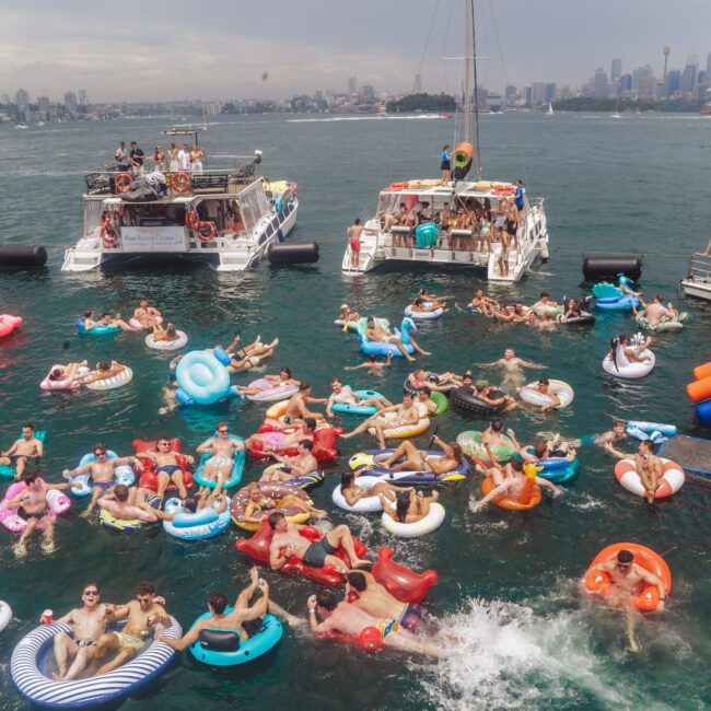 A large group of people relax on colorful inflatable floats in the water near several boats, with a city skyline in the background under a partly cloudy sky.