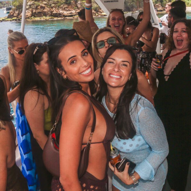 A group of people smiling and posing on a boat during a sunny day, with two women in the foreground looking at the camera and others celebrating in the background. Water and greenery are visible outside.
