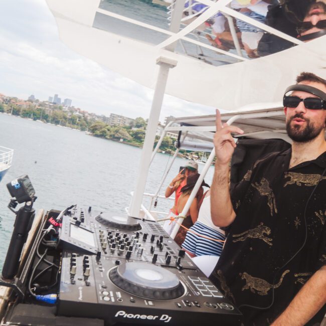 A DJ wearing sunglasses and headphones plays music on a boat deck with DJ equipment. The water and another boat are visible in the background, with city buildings and greenery in the distance.