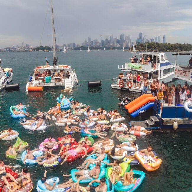 Dozens of people relax on colorful floaties in the water between anchored boats, with city buildings and a cloudy sky visible in the background. The scene is lively and festive.