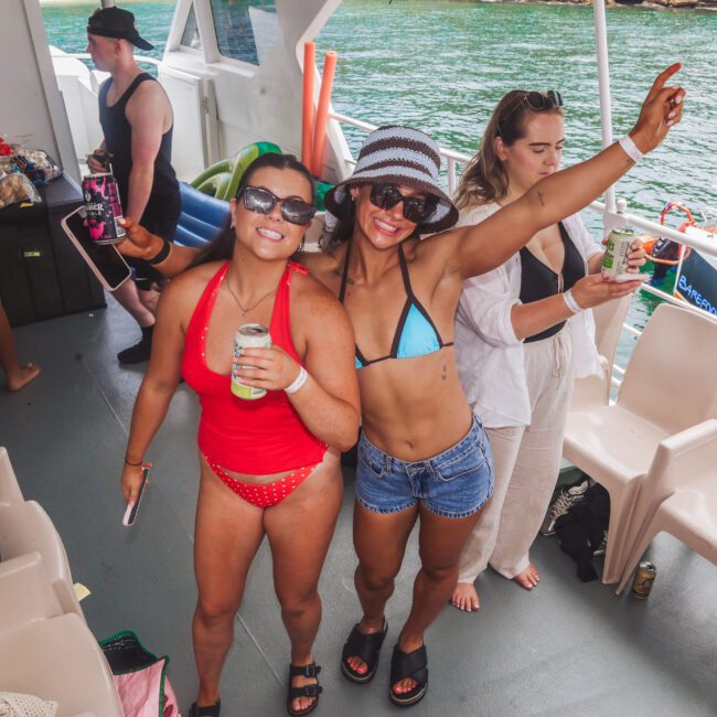 Two smiling women in swimsuits pose with drinks on a boat, surrounded by friends. One woman wears red, the other a blue bikini and hat. Water is visible outside as others mingle and enjoy the party atmosphere.