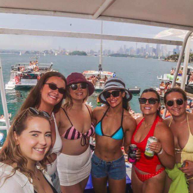 A group of six women in swimsuits, sunglasses, and hats smile for a photo on a boat. Behind them, people relax on boats in the water with a city skyline visible in the background on a sunny day.