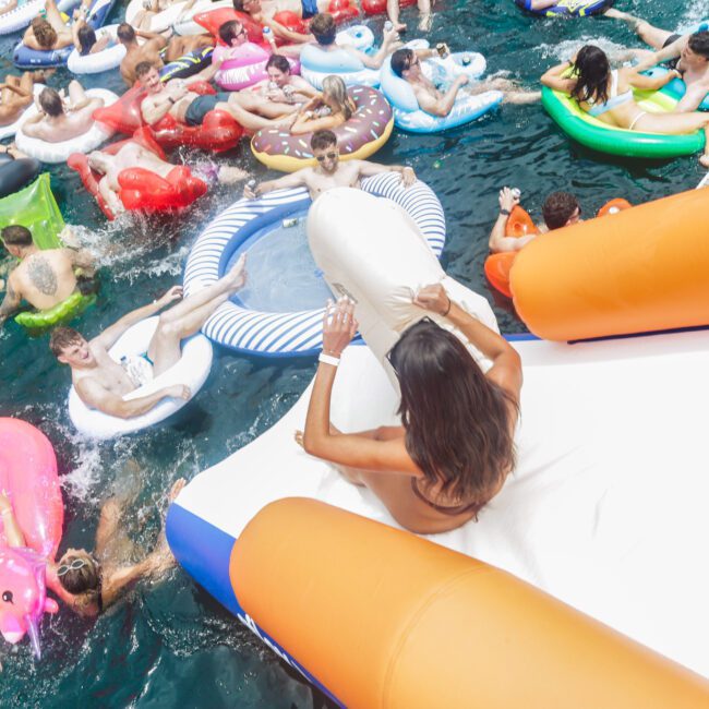 A crowd of people float on colorful inflatable rafts in a pool, while a woman slides down an orange and white inflatable slide toward the water. The scene is bright and lively, with people enjoying a summer day.
