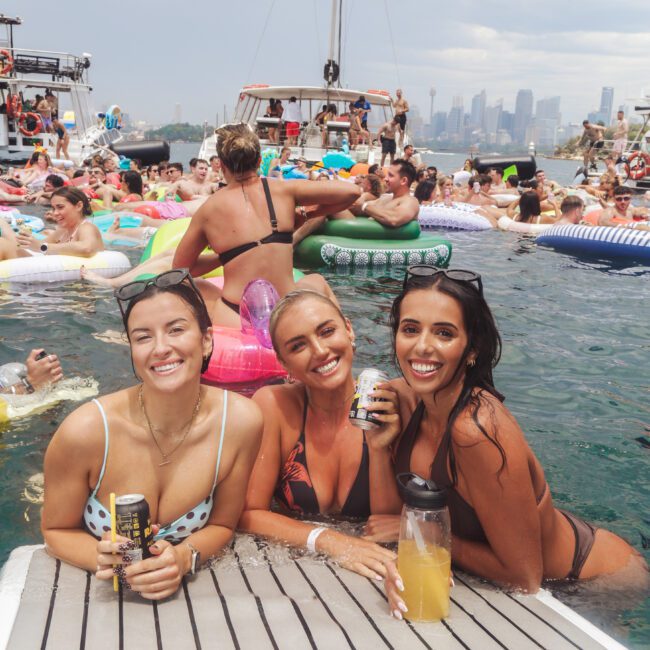 Three women in swimsuits smile and pose with drinks at the edge of a floating dock, surrounded by people on colorful inflatables in the water. Boats and a city skyline are visible in the background under a cloudy sky.