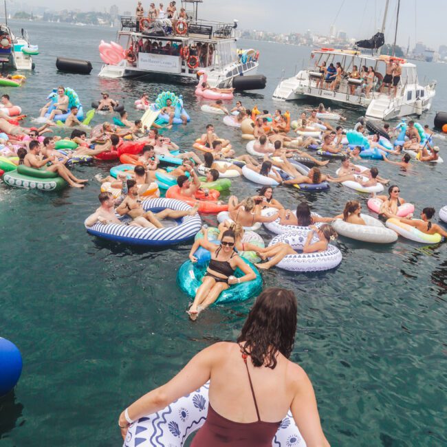 A crowded scene of people lounging on colorful inflatable floats in the water, surrounded by boats with more people on board, all enjoying a lively day under a cloudy sky.