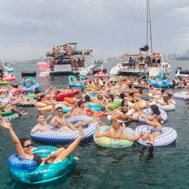 A large group of people float on colorful inflatable tubes in the water, smiling and waving, with boats and a cloudy sky in the background. The scene is lively and festive.