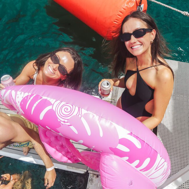 Two women in swimsuits and sunglasses smile on a boat’s metal steps, holding drinks and a pink inflatable float, with turquoise water and people swimming nearby. A red buoy floats in the background.