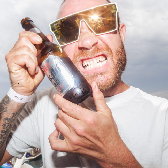 A man in sunglasses grins and holds a beer bottle to his face at an outdoor event. People and boats are visible in the background, and he wears a white wristband and T-shirt. The scene looks festive and lively.