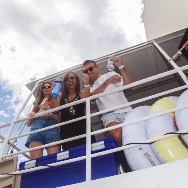 Three people stand on a boat deck holding drinks and smiling, with inflatable pool floats and a blue cooler beside them. The sky is partly cloudy in the background.