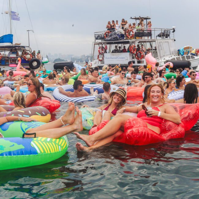 A large group of people enjoy a party on the water, relaxing on colorful inflatable floats near boats. Many are smiling, wearing swimsuits, and holding drinks, with a festive atmosphere under a cloudy sky.