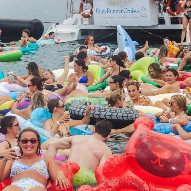A large group of people relaxing and having fun on colorful inflatable floats in the water, with boats in the background. Many are smiling, holding drinks, and enjoying a lively atmosphere under a bright sky.