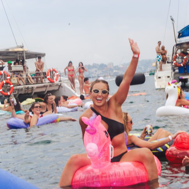 A group of people enjoy a lively party in the water, floating on inflatable pool toys near boats. A smiling woman in sunglasses waves while riding a pink unicorn float. Other people and colorful inflatables surround her.