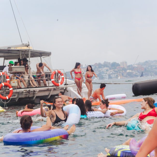 People enjoy a lively day on the water, swimming and floating on colorful inflatables near anchored boats. Some stand on the boats, while others relax in the water. Buildings and hills are visible in the background.